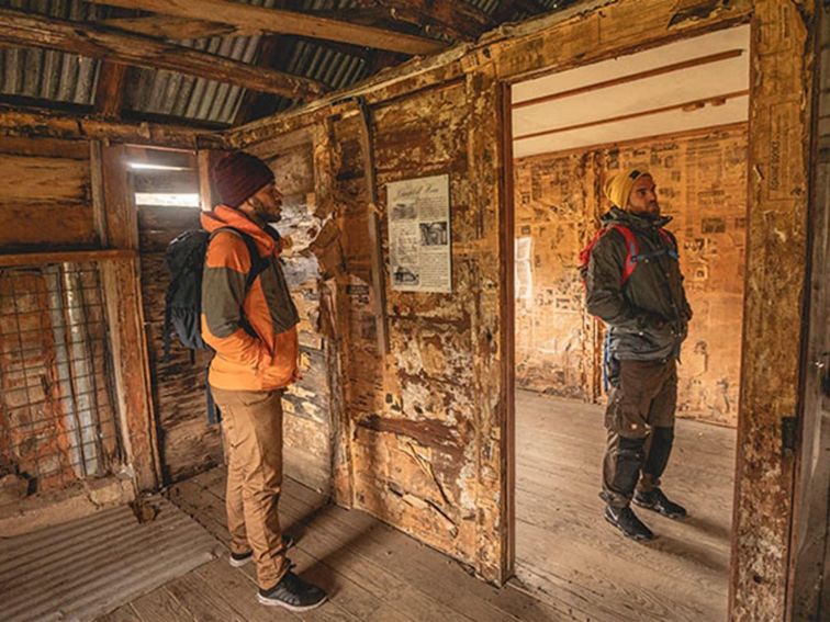 Two men read newspapers lining the walls of a Coolamine Homestead building, Kosciuszko National Park