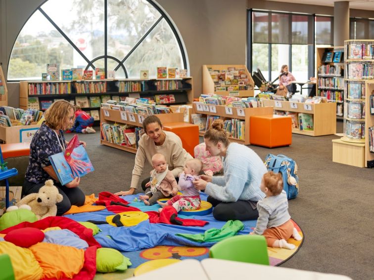 A librarian smiles and reads a book to a small group of babies and mums.