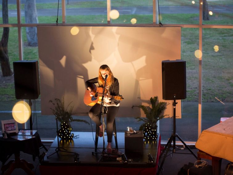 Woman playing a guitar on an informal stage in front of a large window