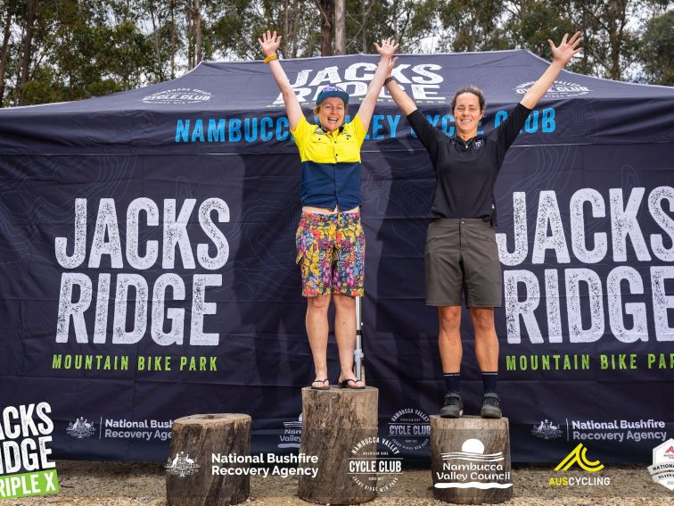 Two ladies on the podium (which are 3 tree trunks) at the XXX, hand in the air in celebration