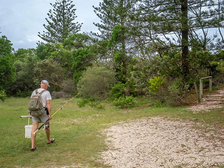 A man with a fishing rod walks across the grass toward a sandy trail and small set of steps. Photo