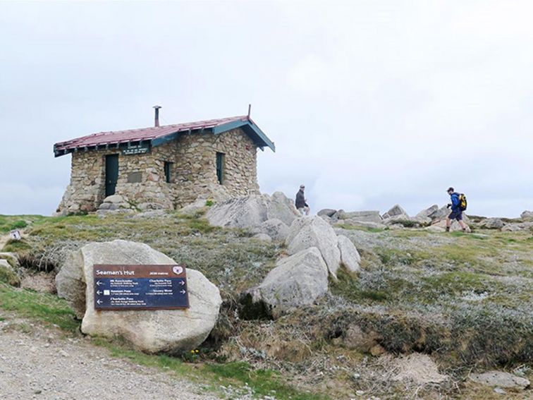 View of hikers passing historic Seamans Hut, with gravel trail and large boulders in the foreground.