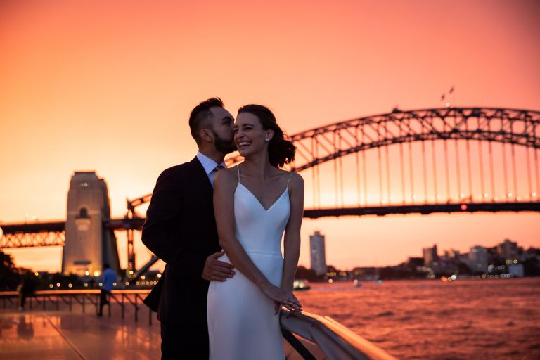 Groom kissing his bride's cheek with Harbour Bridge in background at sunset