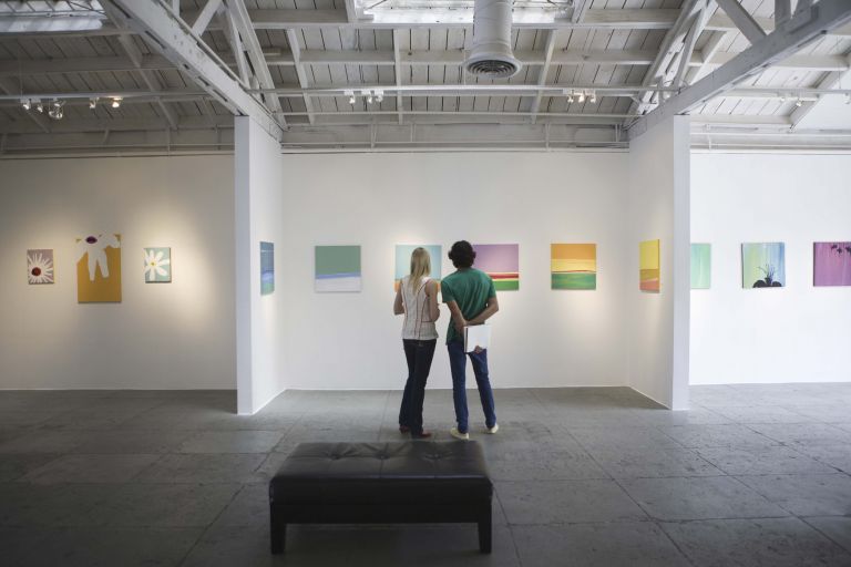 Interior of gallery space with two people looking at artworks on back wall, ceiling with lighting and aircon visible above.