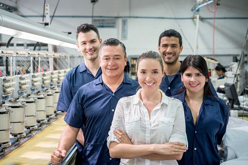Group of workers and trainees smiling and standing in row besides textile factory machinery