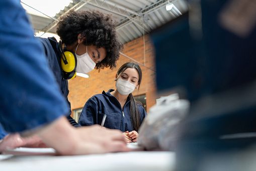 Two apprentices in masks in a factory leaning over a drafting table