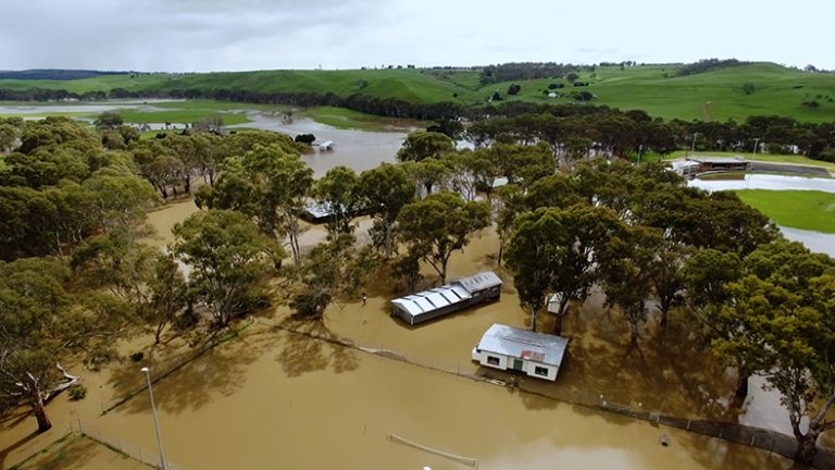 Aerial view of a flooded farm showing a home, sheds and trees sitting in brown water, with green hills in the background