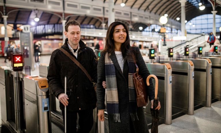 Man with white stick holding arm of woman at ticket barrier in train station