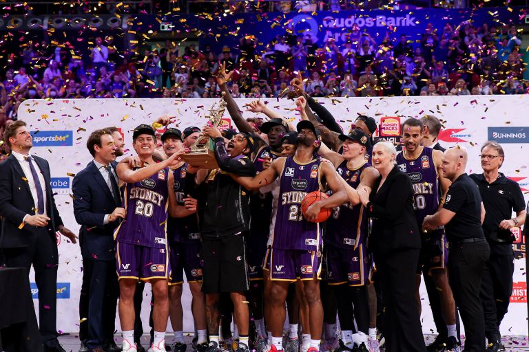 The Sydney Kings basketball team are standing on a stage. Three players are holding a large trophy up high celebrating and the rest of the team have their arms up celebrating with them. There is confetti falling around them.