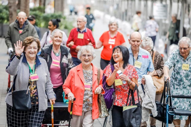 Friends and happy faces in crowd of people walking along street towards Seniors Festival event