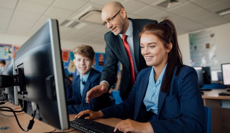 Two pupils, a male and a female, are sitting behind computers receiving help from a male teacher during a lesson.
