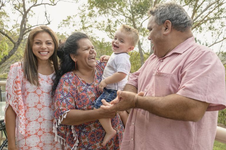 An Aboriginal family is standing in front of the camera. Four people are in the image, with the central figure being a young boy who is being held in his grand mother's arms. The family are all laughing at the young child.