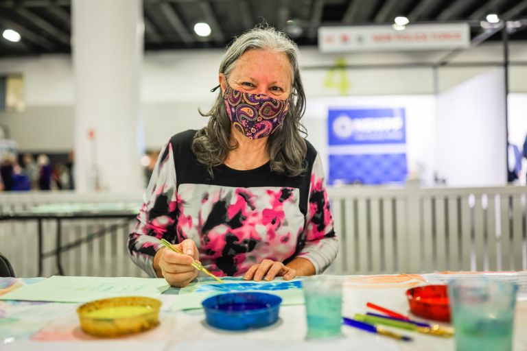 Lady smiling beneath face mask, as she paints art at seniors expo activity
