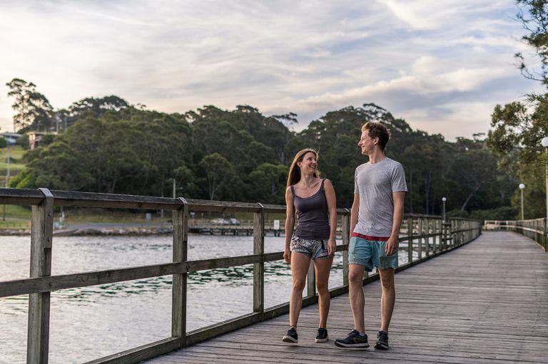 A young man and woman smiling, walking along the Mill Bay Boardwalk, Narooma NSW