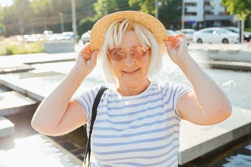 Seniors woman stands against urban waterside, clutching hat and wearing sunglasses.