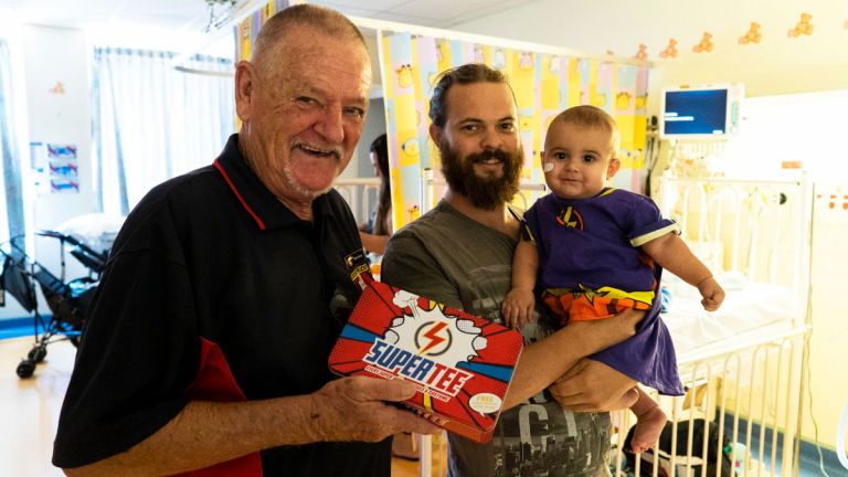 A senior man gives a donation to a dad and his infant child inside a hospital ward, they are all looking at the camera smiling