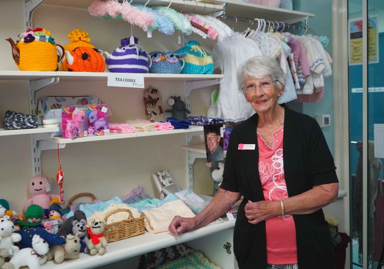 A gift shop volunteer stands by a shelf of knitted tea cosies and handmade toys. She smiles at the camera