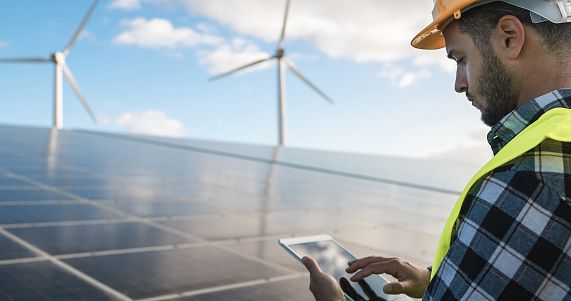 Worker in PPE glances down at ipad with solar panels and wind turbine in background