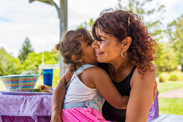 Photo of Aboriginal grandmother receiving a kiss on the cheek from her granddaughter. The grandmother has brown curly hair and black sunglasses sitting on her head. The granddaughter is sitting on her lap hugging her, she's wearing a pink skirt and white singlet. They are sitting in a park and the park table, with food on top, and green trees, are in the background. 