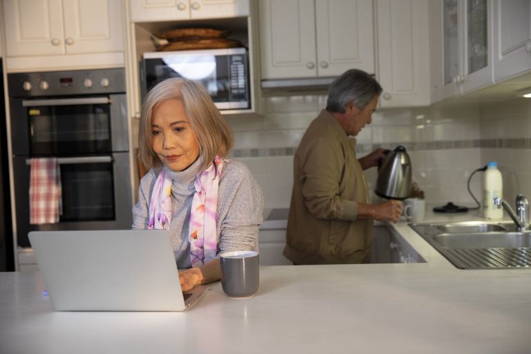 A woman on laptop in kitchen. A man is pouring a cup of tea in the background.