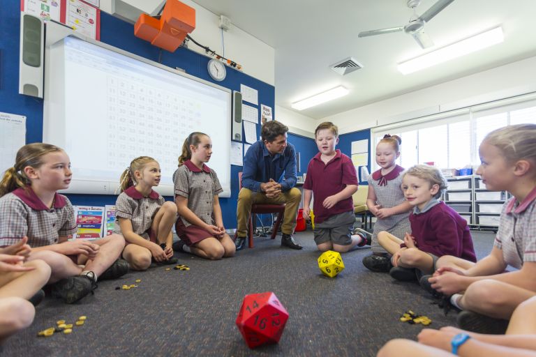 A school teacher and primary school students sit in a circle playing with a ball inside a classroom.