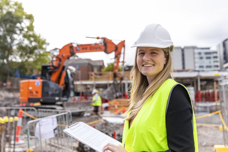 Stephanie works as a Project Manager for Turner and Townsend at the North Sydney Public School redevelopment. She is standing on site in hi-vis and a helmet, smiling at the camera with a bulldozer in the background.
