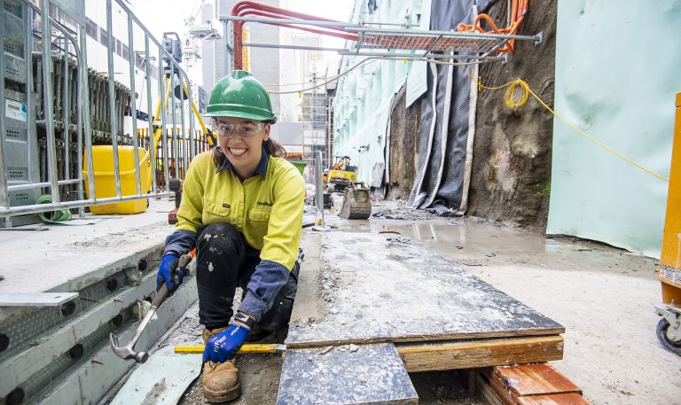 Alyssa works as a builder for Lendlease at the Victoria Cross Station. She is bent down with a hammer and chisel on a worksite.  