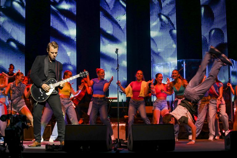 Photo of a bustling stage with Rodger Corser on the left playing a guitar; a band and dancers in the middle and someone doing a cartwheel on the left. 