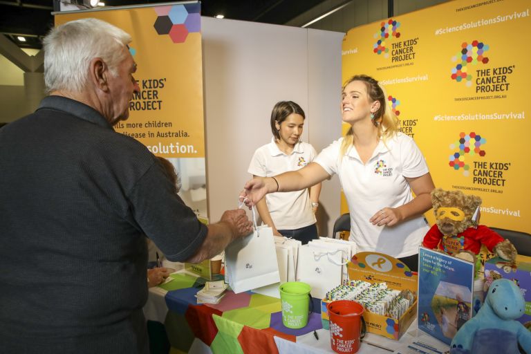 woman at kids cancer project expo stall handing over showbag to male patron