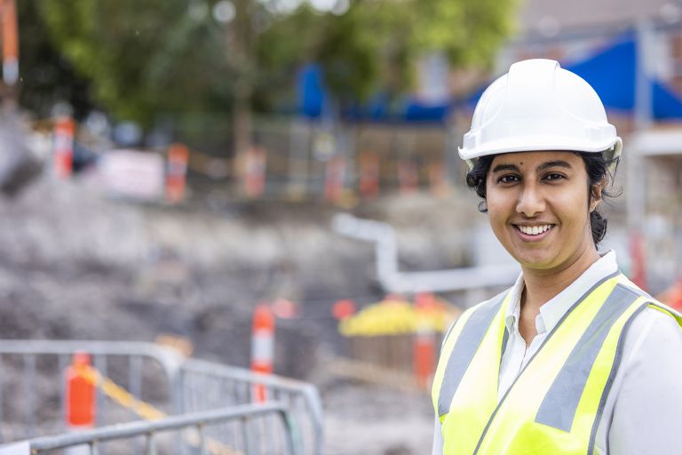 Tarini works as a Project Manager for Turner and Townsend at the North Sydney Public School redevelopment. She is standing in front of a mound of dirt, in front of a barricade wearing hi-vis and a hard hat.