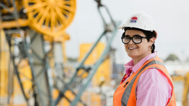 Teresa is standing in front of large machinery wearing a pink t-shirt, hi-vis vest and hard hat.