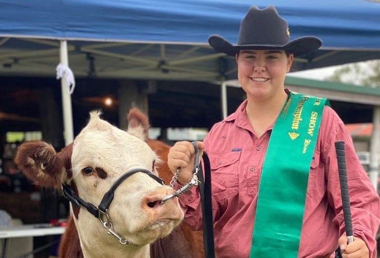 Woman on the right wearing a pink shirt, hat and a green sash for women. A cow stands next to her.