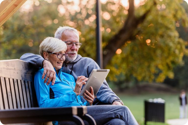 Two seniors looking at an ipad on a park bench outside