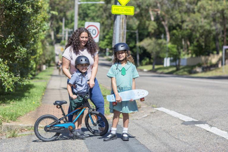 A mother with two children waiting to cross the road together. The children are wearing helmets, with one holding a skateboard and the other with a bicycle.