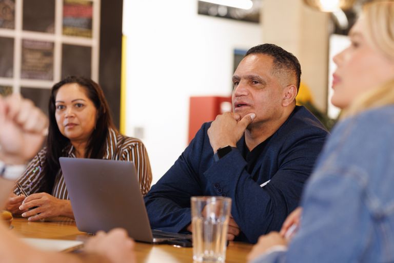 An Aboriginal man is sitting at a conference table leading a meeting. He is wearing a navy blue shirt and has a laptop in front of him.
