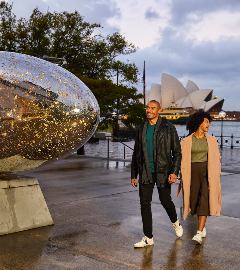 Picture of a couple, walking holding hands with the Sydney Opera house behind them, next to a large reflective scultupture