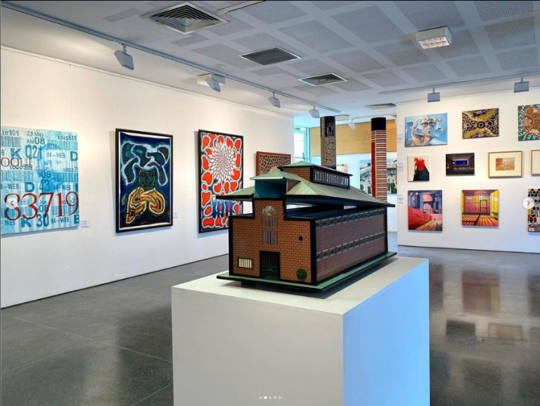 Interior of art gallery exhibition space with model of building on white plinth in foreground and artwork on walls.