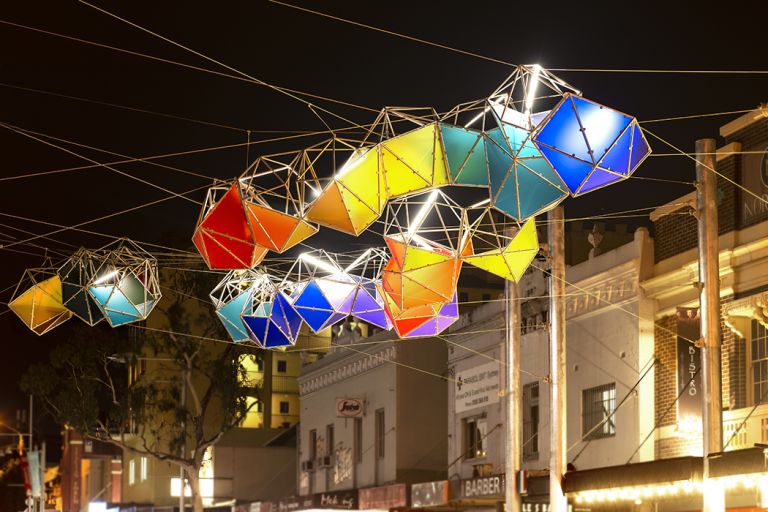 Colourful box-kite-like sculpture lamps hanging over street of terrace houses at night