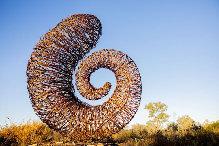 A snail shell like spiral sculpture, made out of steel, freestanding in an outdoor landscape against a blue sky.