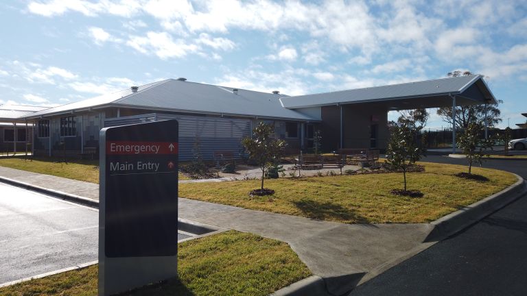 Main entrance to the Murrumburrah-Harden Health Service. A sign indicating the main and emergency entry points is shown to the left of the image. A curved bitumen driveway leads to the entry points. A part of a car park is shown on the left of the image. A concrete walkway runs along the topside of the car park. 