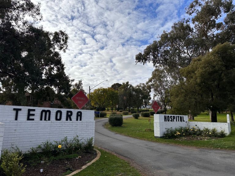 The main entry point to the Temora Hospital. Two white brick walls flank the main driveway. The left white wall has 'Temora' lettered in black, the right white wall has 'Hospital' lettered in black. Each wall also features a red diamond-shaped sign that says 'Entrance Only'. The driveway leads up the main hospital building.