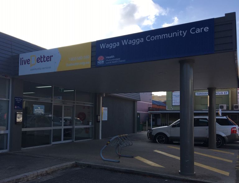Main entrance to the Wagga Wagga Community Care centre. There is a bicycle ramp for 4 bicycles outside the main entrance. To the right, there are a couple of car parking spaces. Along the top left of the building's entrance awning the centre's signage shows 'live Better community living' and 'Wagga Wagga Community Care'.  