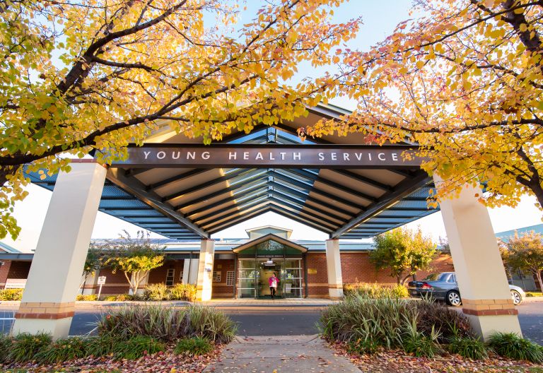 A photo of the entrance of Young Health Services, a modern red brick building with a blue roof and white walls, surrounded by trees with yellow leaves.
