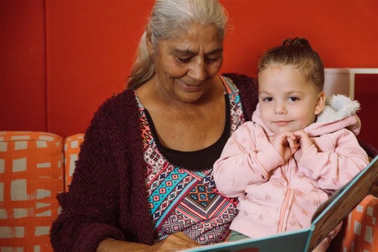 Image of a woman reading to her grandchild. The grandmother is wearing a brown cardigan and colourful top and is holding a book. The granddaughter is sitting on her lap wearing a pink hooded sweatshirt with her hands underneath her chin. 