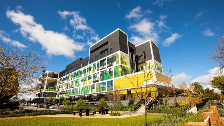 Exterior view of the Wagga Wagga Health Service building. The building features shades of green and yellow clad ties along the bottom and middle and black cladding at the top. A ramp is shown to the far right of the image. Steps and outdoor seating among landscaped green spaces is shown in the foreground of the image. To the left, car parking spaces sit in front of the main entrance. 