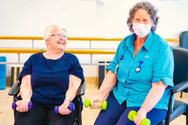 An older female resident holds dumbbells and smiles as she works with a female healthcare worker