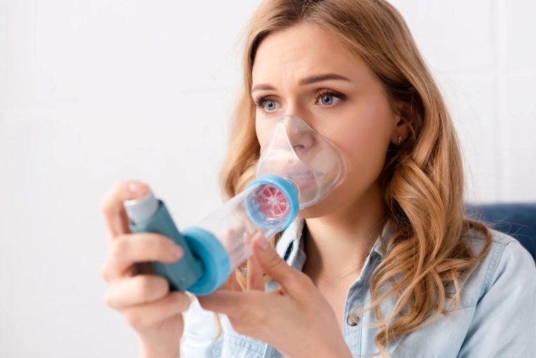 A woman facing toward the left uses an asthma puffer and inhalation device.