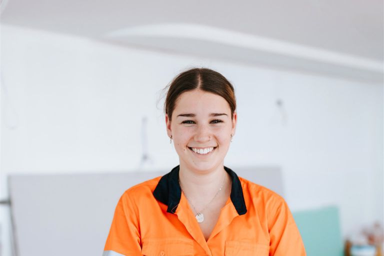 Ashlyn is standing in front of a white wall, wearing hi-vis, smiling. 