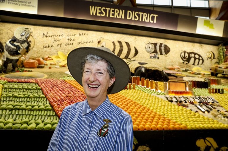 Smiling volunteer at the Easter Show, Western Districts Exhibit. The volunteer is wearing a blue and white striped shirt with badge, and a hat. They stand in front of a display of fruits and food in jars. 