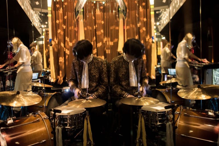 A drumer in a leopard print jackets playing in front of a mirro giving the illusion there are two people. 